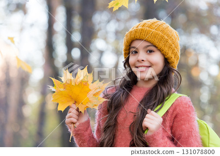 smiling teen child in knitted hat and sweater carry school backpack hold yellow maple leaves in forest of park in autumn season with warm weather, back to school 131078800