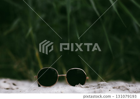 Stylish round sunglasses on sand, with a blurred green background. A pair of round sunglasses with black glasses with green foliage in the background. 131079193