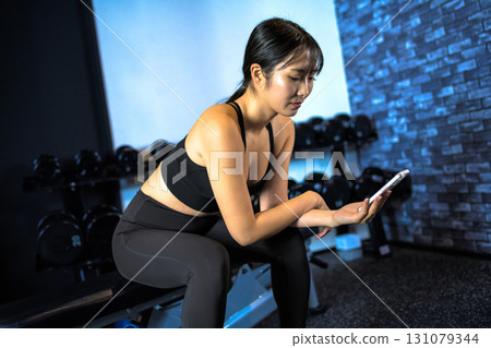 A woman taking a break while sitting and looking at her smartphone during cool time. A cool woman exercising at the gym. A woman doing muscle training. A woman taking a break while sitting and looking at her smartphone during cool time. A cool woman exercising at the gym. A woman doing muscle training. 131079344
