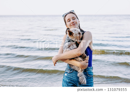A young pretty brunette woman cuddles adorable Yorkshire terrier doggy on a sunny beach day at the sea. A girl smiles while holding a dog at the shore A young pretty brunette woman cuddles adorable Yorkshire terrier doggy on a sunny beach day at the sea. A girl smiles while holding a dog at the shore 131079355