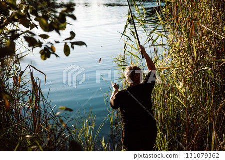 A mature woman fishing on a sunny day on a lake shore. A back view of a person fishing on a river bank, coast. A mature woman fishing on a sunny day on a lake shore. A back view of a person fishing on a river bank, coast. 131079362
