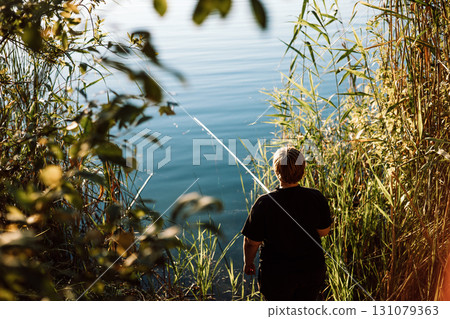 A mature woman fishing on a sunny day on a lake shore. A back view of a person fishing on a river bank, coast. A mature woman fishing on a sunny day on a lake shore. A back view of a person fishing on a river bank, coast. 131079363