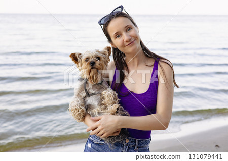 Happy young woman in swimsuit holding small dog at the beach. Authentic summer portrait with real emotions, Perfect for pet and travel themes. 131079441