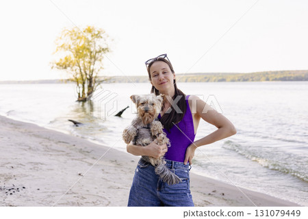 Happy young woman in swimsuit holding small dog at the beach. Authentic summer portrait with real emotions, Perfect for pet and travel themes. 131079449