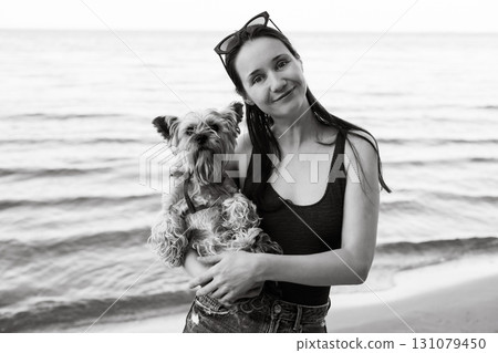 A smiled and relaxed young woman holding her small dog enjoy a day at the beach on summer vacation. A woman holds a Yorkshire terrier doggy on hands 131079450