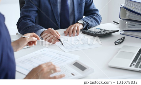 Accountants analyzing financial documents, calculating numbers together at shared workspace with laptop and office supplies surrounding them. Audit and taxes in business Accountants analyzing financial documents, calculating numbers together at shared workspace with laptop and office supplies surrounding them. Audit and taxes in business 131079832