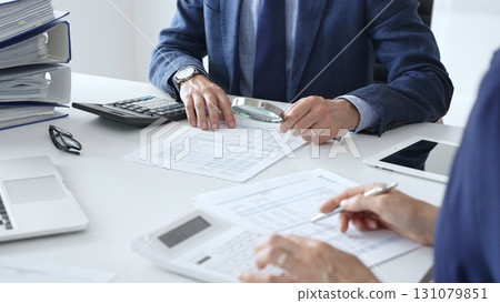 Close up of male financial analysts with female colleague examining documents, using magnifying glass and calculator at workplace in office. Audit and taxes in business 131079851