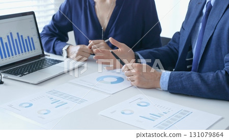 Businessman and businesswoman are analyzing financial data on a laptop and printed documents, pointing at charts and graphs during a meeting in bright office. Business people and finance concept 131079864