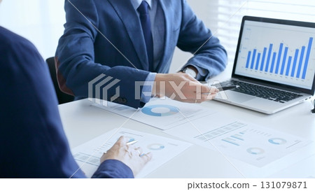 Business colleagues discussing financial data on a laptop and printed documents while analyzing charts and market trends. Business people and finance concept Business colleagues discussing financial data on a laptop and printed documents while analyzing charts and market trends. Business people and finance concept 131079871