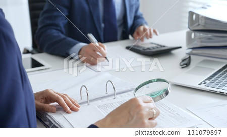 Business colleagues are working together, analyzing financial documents with a magnifying glass, calculating and taking notes in the office. Audit and taxes in business Business colleagues are working together, analyzing financial documents with a magnifying glass, calculating and taking notes in the office. Audit and taxes in business 131079897