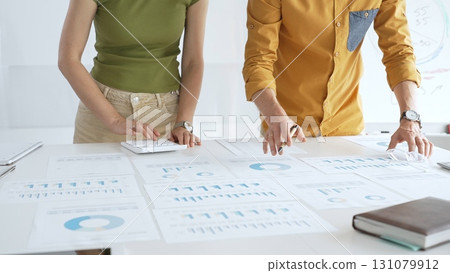 Casual business people are working together, analyzing financial data and reports using a keyboard and printed charts spread out on a table in a modern office 131079912