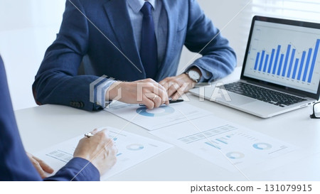 Business colleagues discussing financial data on a laptop and printed documents while analyzing charts and market trends. Business people and finance concept Business colleagues discussing financial data on a laptop and printed documents while analyzing charts and market trends. Business people and finance concept 131079915