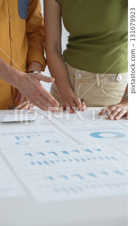Business professionals reviewing financial charts, calculating data with calculator on table, analyzing statistics during collaborative project meeting, close up vertical view Business professionals reviewing financial charts, calculating data with calculator on table, analyzing statistics during collaborative project meeting, close up vertical view 131079923