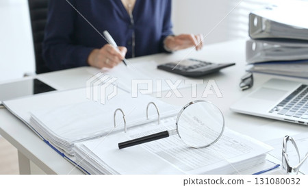 Magnifying glass and calculator are on the table over financial documents opposite female accountant analyzing data, reviewing reports, binders and laptop on a white desk. Audit and taxes concept 131080032