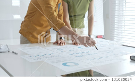 Two business professionals are examining financial reports, using a pen to point at specific data points on charts and graphs spread out on a white desk Two business professionals are examining financial reports, using a pen to point at specific data points on charts and graphs spread out on a white desk 131080034