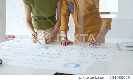 Two business professionals are examining financial data and reports, pointing at charts and graphs displayed on a table during a collaborative office meeting 131080039