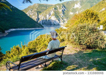 Wide shot of a young man wearing a hat sitting on a bench, enjoying the panoramic view of mountains and Piva Lake in Montenegro. Travel, nature, and lifestyle concept. Wide shot of a young man wearing a hat sitting on a bench, enjoying the panoramic view of mountains and Piva Lake in Montenegro. Travel, nature, and lifestyle concept. 131080048