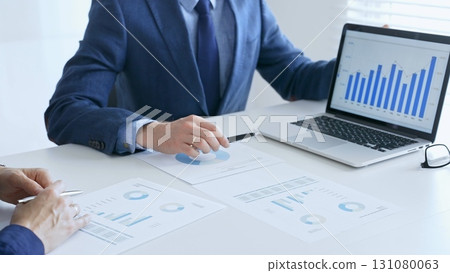 Business colleagues discussing financial data on a laptop and printed documents while analyzing charts and market trends. Business people and finance concept Business colleagues discussing financial data on a laptop and printed documents while analyzing charts and market trends. Business people and finance concept 131080063
