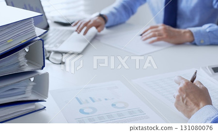 Team of accountants or auditors are analyzing financial reports and making notes while sitting at a white table, close up of a stack of blue folders full of paperwork. Audit and taxes theme in 131080076