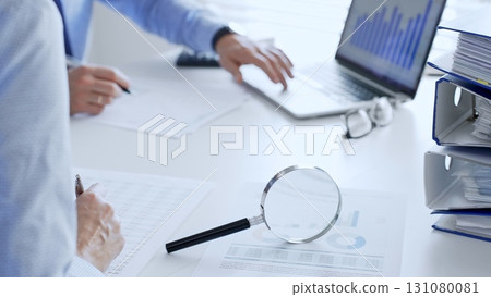 Professional colleagues reviewing financial reports, examining graphs and documents with magnifying glass, laptop on minimalist white workspace. Audit and taxes in business 131080081