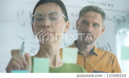 Professional woman with glasses marking glass board, arranging sticky notes while male colleague reviews strategic planning within modern, well lit workspace. Business people concept 131080328