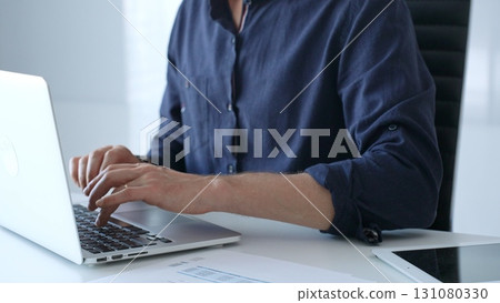 Businessman wearing a blue shirt is typing on a laptop with financial reports and a tablet on his desk, working diligently in a modern office environment. Business people concept Businessman wearing a blue shirt is typing on a laptop with financial reports and a tablet on his desk, working diligently in a modern office environment. Business people concept 131080330