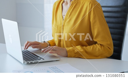 Professional female executive wearing yellow blouse working on laptop at minimalist white desk in contemporary workplace, typing with focus. Business people concept Professional female executive wearing yellow blouse working on laptop at minimalist white desk in contemporary workplace, typing with focus. Business people concept 131080351