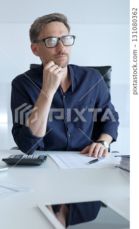 Businessman sitting at a sleek desk, working on a laptop and thinking in a bright, modern office filled with natural light. Business people concept Businessman sitting at a sleek desk, working on a laptop and thinking in a bright, modern office filled with natural light. Business people concept 131080362