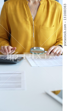 Female accountant wearing yellow shirt is examining a financial report with a magnifying glass while using a calculator and laptop in an office setting, close up. Audit and taxes in business Female accountant wearing yellow shirt is examining a financial report with a magnifying glass while using a calculator and laptop in an office setting, close up. Audit and taxes in business 131080363