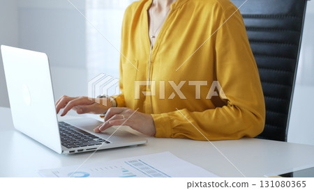 Unknown businesswoman wearing yellow blouse working on laptop at minimalist white desk in contemporary workplace, typing with focus. Business people concept 131080365