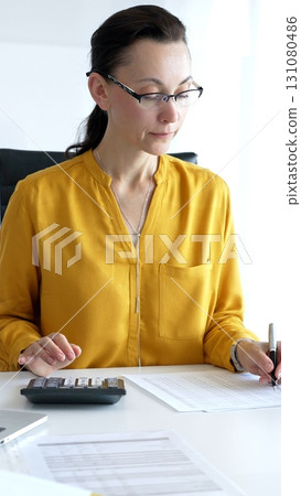 Businesswoman wearing yellow shirt is sitting at white desk and using calculator while taking notes, with laptop and folders in the background. Audit and taxes in business 131080486