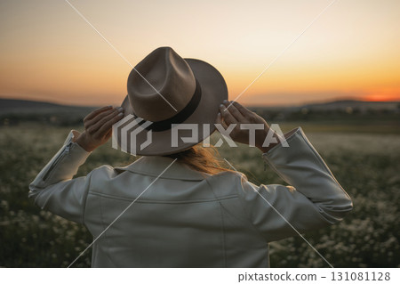 A woman wearing a hat and a white jacket stands in a field. The woman is looking at the camera. A woman wearing a hat and a white jacket stands in a field. The woman is looking at the camera. 131081128