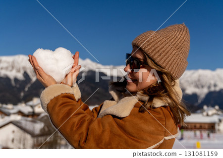 Snow Heart Mountains Winter: Woman holding snow heart, Alpine village backdrop, sunny winter day, romantic gesture. 131081159