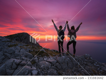 Women with backpacks and raising arms on mountain at sunset Women with backpacks and raising arms on mountain at sunset 131081642
