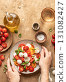 Caprese salad with sliced tomatoes, mozzarella, basil, olive oil, hands holding on brown textured background. Top view 131082427