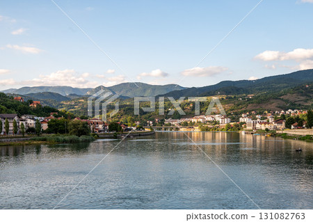 A small coastal town with houses and buildings, located by a calm river, surrounded by wooded hills A small coastal town with houses and buildings, located by a calm river, surrounded by wooded hills 131082763