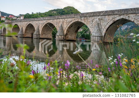 An ancient stone bridge with arches spanning the tranquil Drina River with wild flowers 131082765