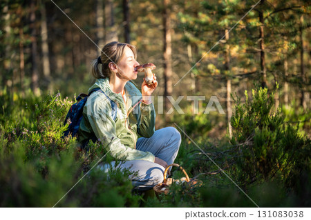 Content woman forager holding boletus mushroom and smelling scent, sit in heather bushes in woodland 131083038