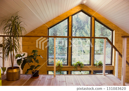 A cozy wooden cabin loft with a large window overlooking a lush green forest. Interior view of a wooden cabin loft, a large window with a forest view, A cozy wooden cabin loft with a large window overlooking a lush green forest. Interior view of a wooden cabin loft, a large window with a forest view, 131083620