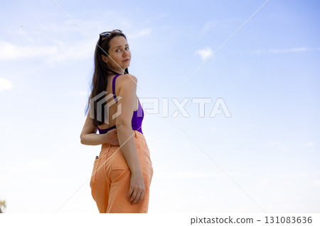 Trendy young woman in colorful outfit posing outdoors against blue sky. Serene girl in orange pants and swimsuit looks back at the camera. 131083636