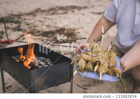Preparing marinated chicken skewers over a charcoal grill on the beach A person prepares marinated chicken skewers for grilling over a charcoal fire on the beach, setting up for a summer cookout. 131083854