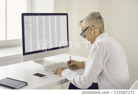Senior man taking notes on paper while sitting at office desk with desktop computer 131087171