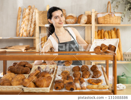 Young woman employee puts croissants in window, arranges display of goods at bakery. 131088886