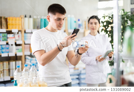 Guy in pharmacy scans QR code on antifungal ointment, pharmacist in background 131089002