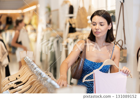 Girl customer chooses tank top in store, and buyer carefully examines product. Girl customer chooses tank top in store, and buyer carefully examines product. 131089152