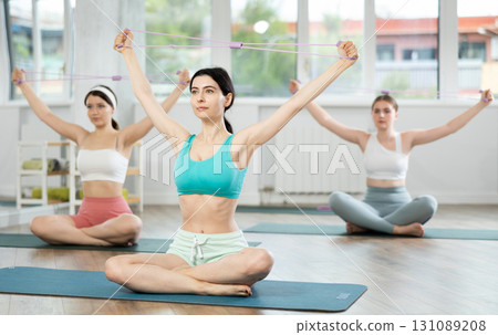 Sitting on mats in Pilates studio, young girls do arm stretches using an expander Sitting on mats in Pilates studio, young girls do arm stretches using an expander 131089208