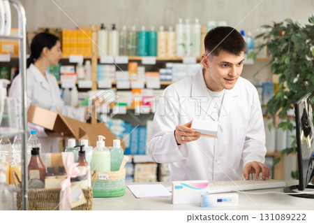 Portrait of young guy pharmacist standing at prescription counter and working on computer, stocktaking medicines at pharmacy shop and female assistant in background Portrait of young guy pharmacist standing at prescription counter and working on computer, stocktaking medicines at pharmacy shop and female assistant in background 131089222