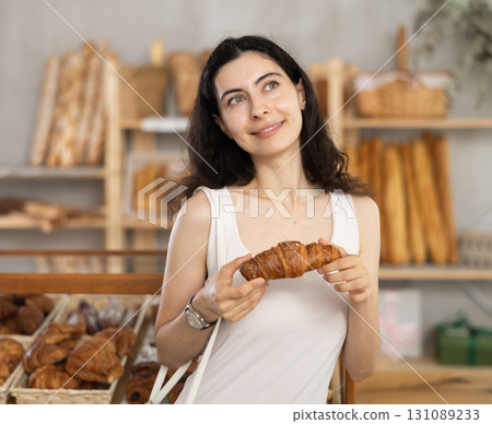 Young female customer stands with croissant in hands near window of bakery. 131089233