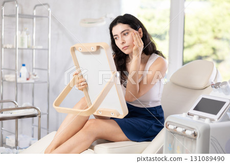 Young brunette woman admires her new face in mirror after plastic surgery 131089490