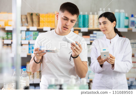 Young female pharmacist helping young man choosing skin care lotion or cream in drugstore 131089503